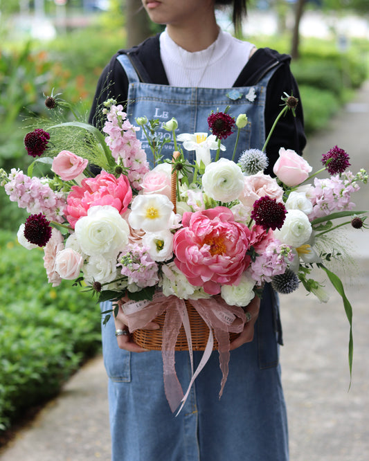 A person holding a basket of assorted flowers, predominantly pink peonies, tied with a ribbon.