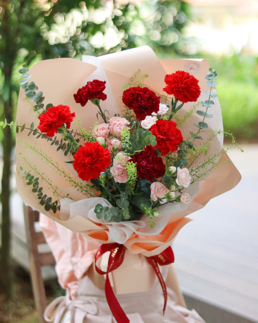 Bouquet of red and pink flowers wrapped in paper with a red ribbon, set against a blurred natural background.